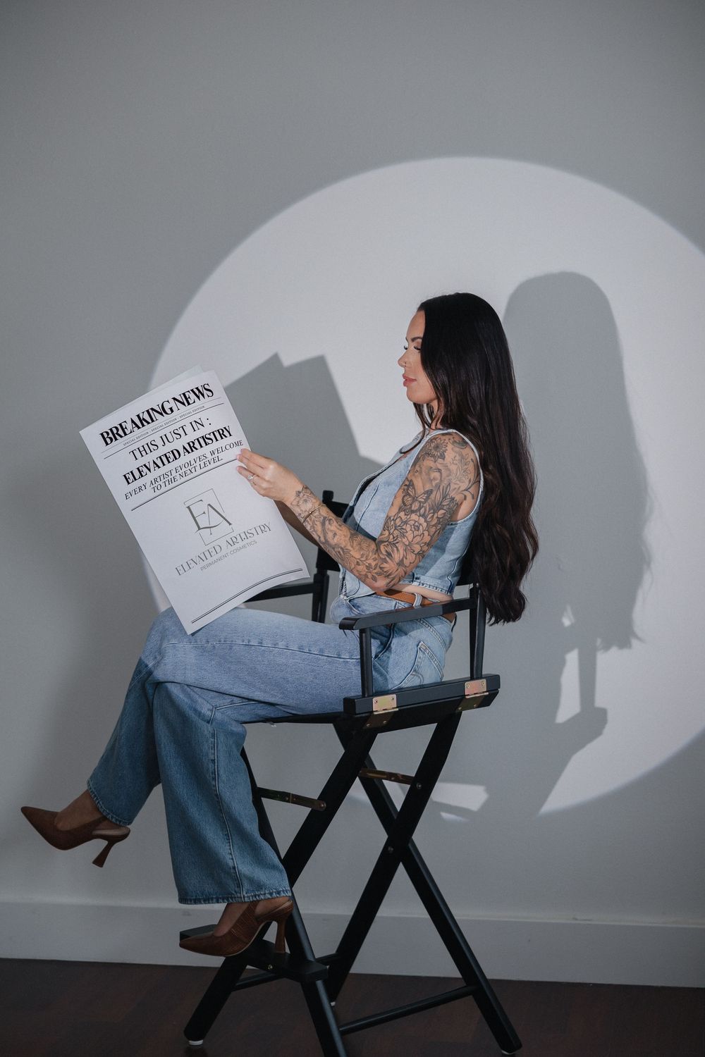 A person in jeans and brown boots sits in a director's chair reading a document, casting a dramatic shadow on a gray wall.