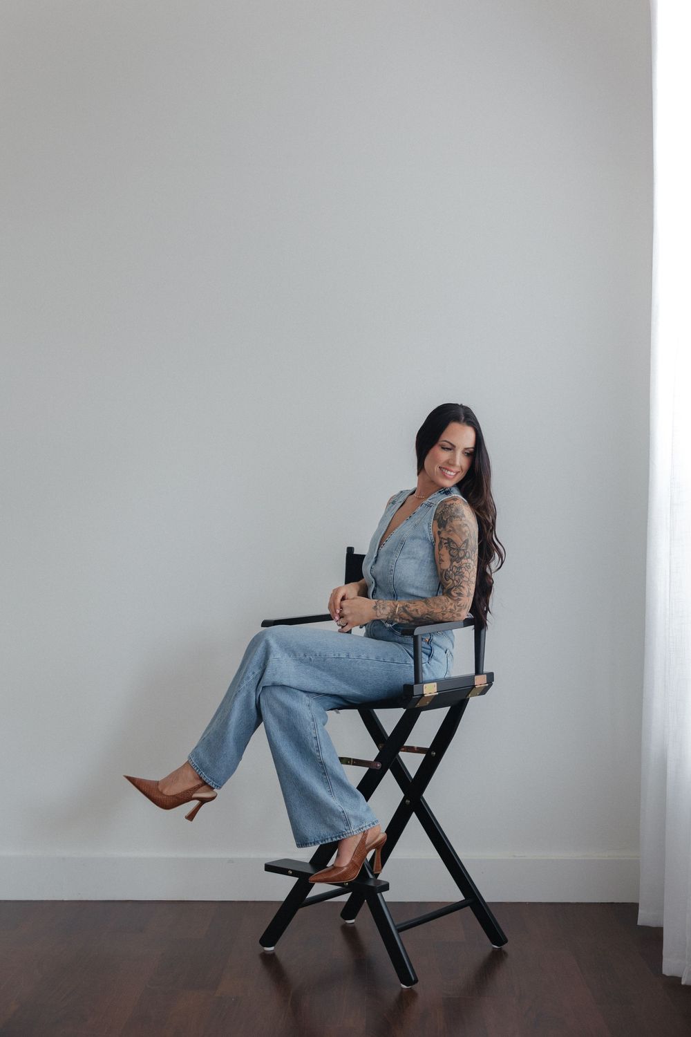 Woman in denim overalls and sleeve tattoos poses on a black director's chair against a white wall.