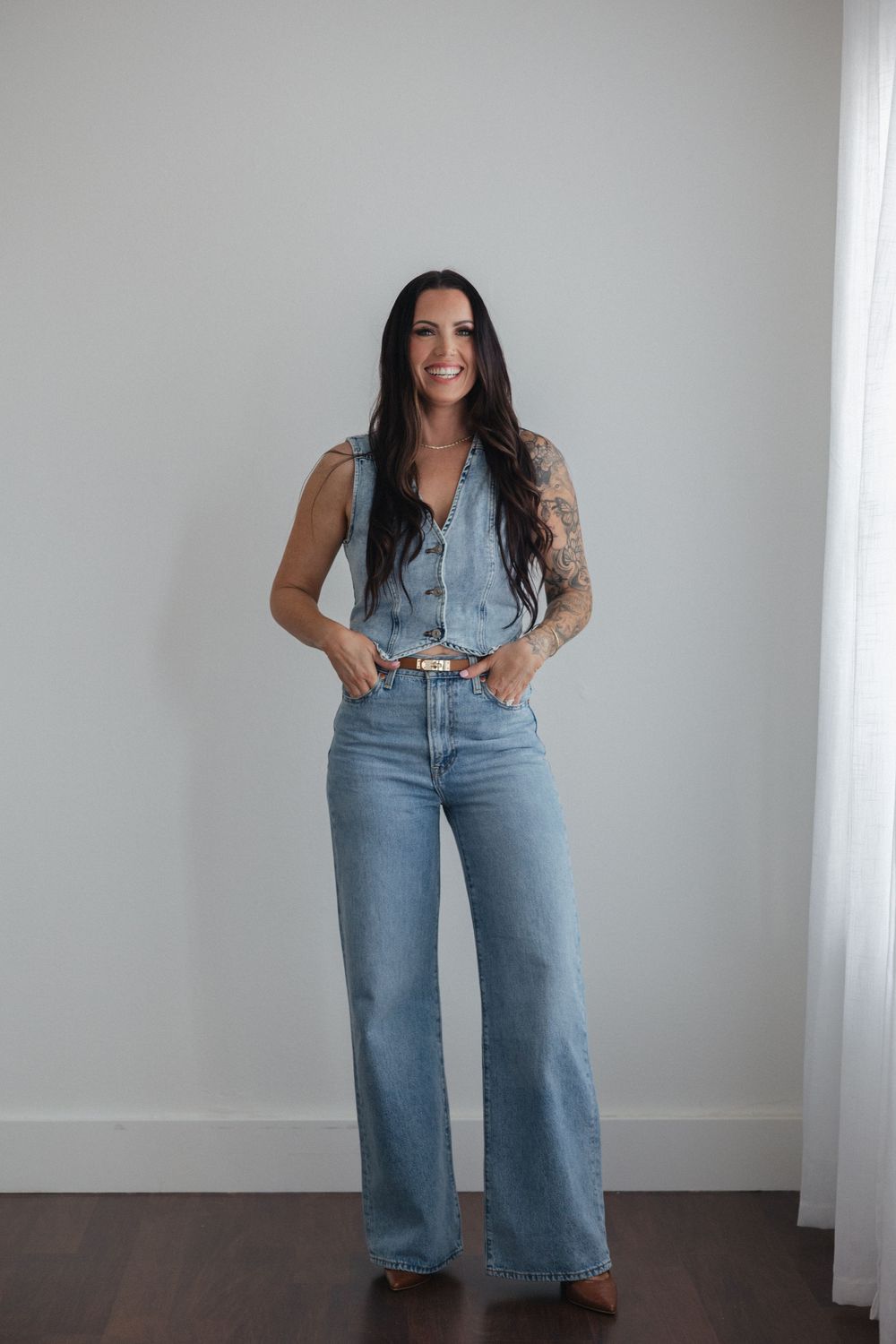 A casual denim outfit featuring wide-leg jeans and a sleeveless button-up vest against a white wall interior.