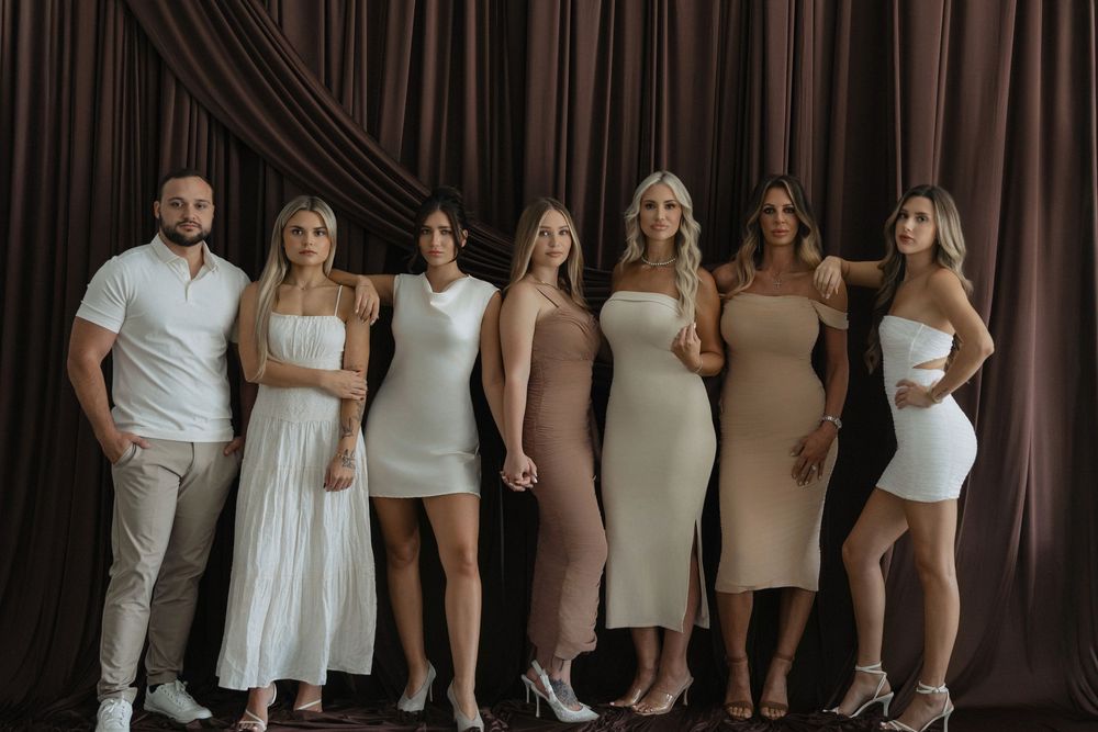 Group of seven people posing together in coordinated white and nude outfits against a dark brown draped backdrop.