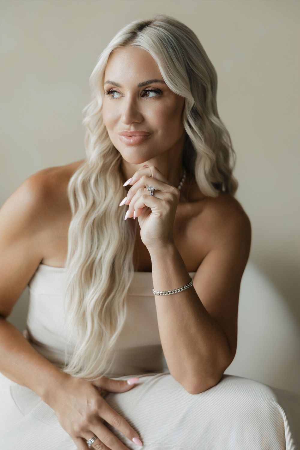 Elegant blonde woman in white strapless dress with diamond jewelry, posing gracefully in a bright, neutral-toned setting.