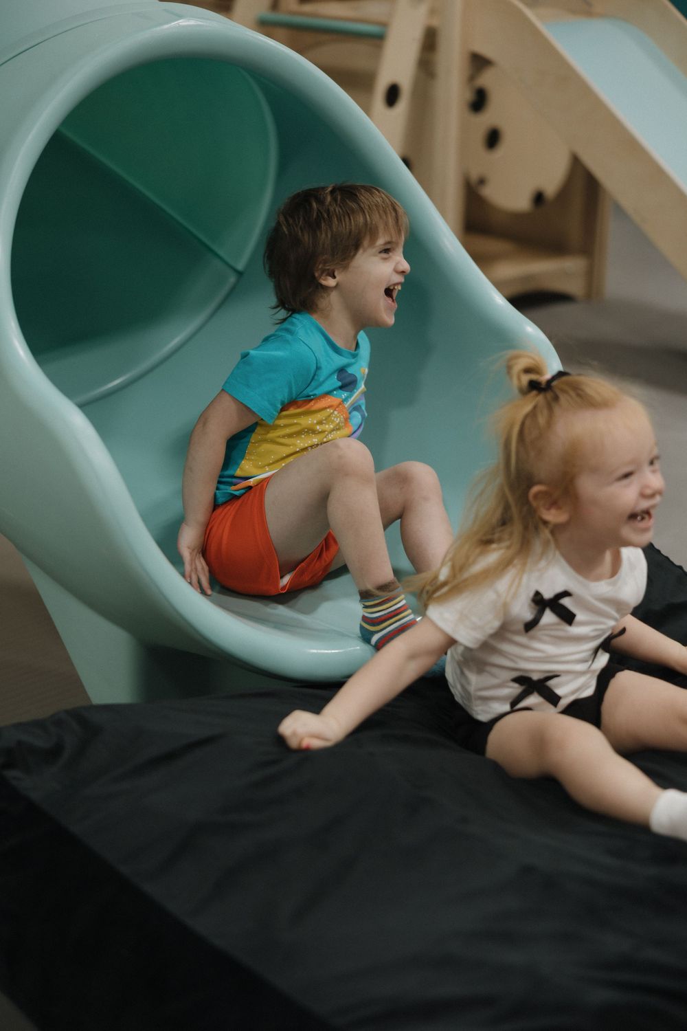 Two happy children laughing and playing on a teal tunnel slide at an indoor playground.