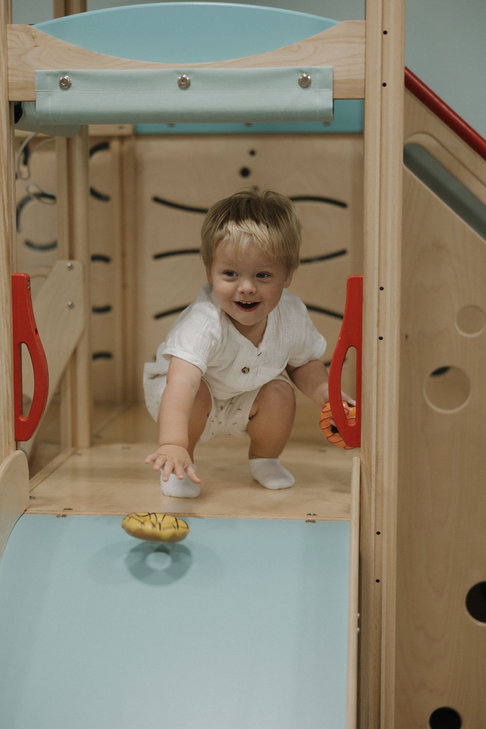 Smiling toddler playing on a wooden indoor playground climbing structure with blue accents and red handles.