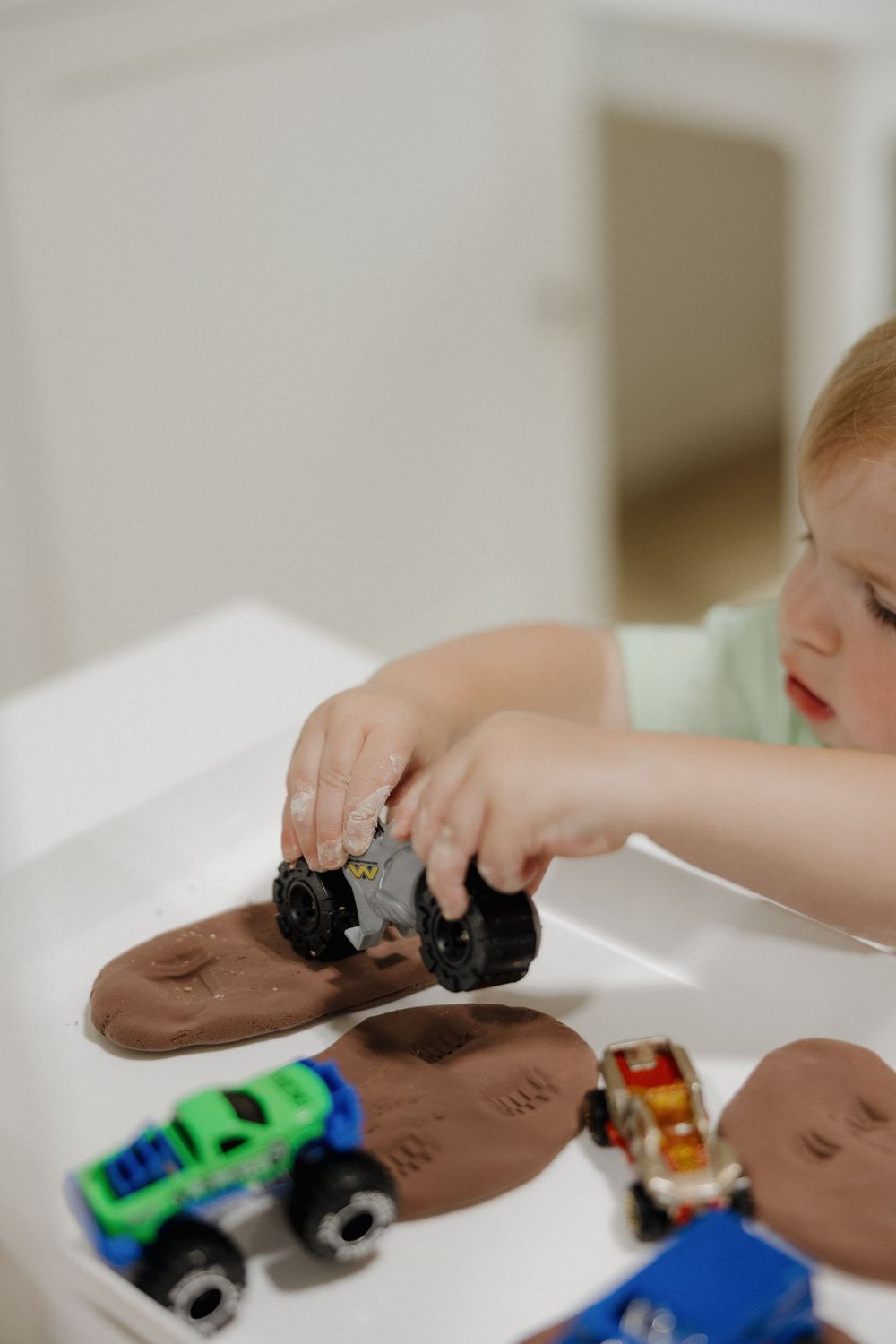 Young toddler pressing toy monster trucks into brown play dough on a white table, creating tire track impressions.