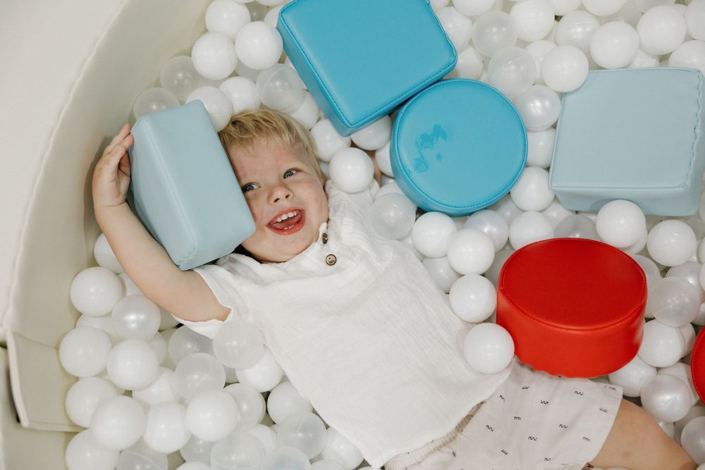 Happy toddler playing in white ball pit with colorful plastic containers, smiling and holding blue box.