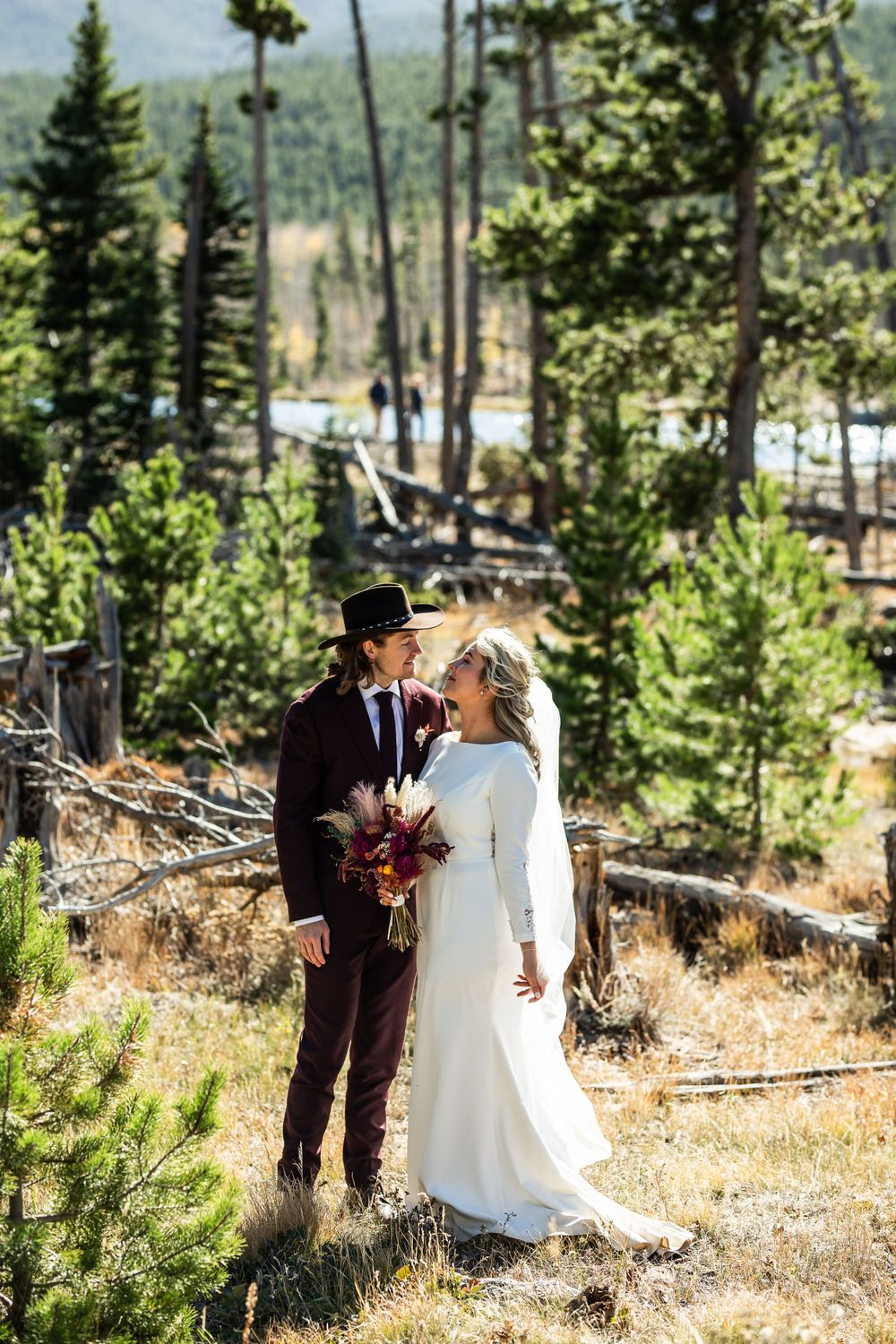 A wedding couple stands in a scenic mountain forest setting at altitude with evergreen trees and distant views.