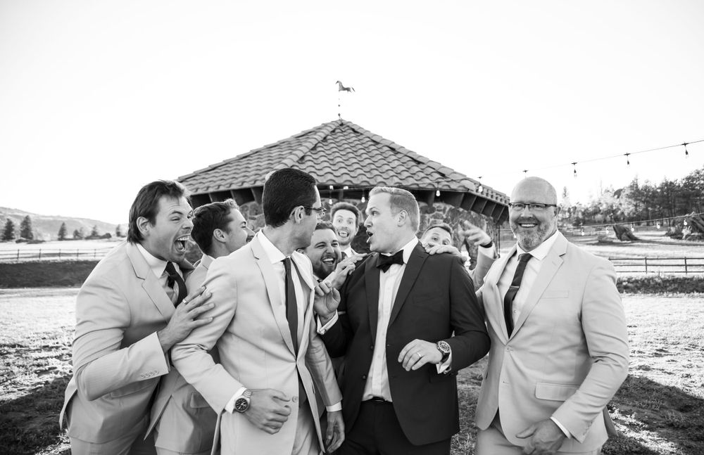 Black and white photo of groomsmen sharing a laugh outside a rustic stone gazebo.