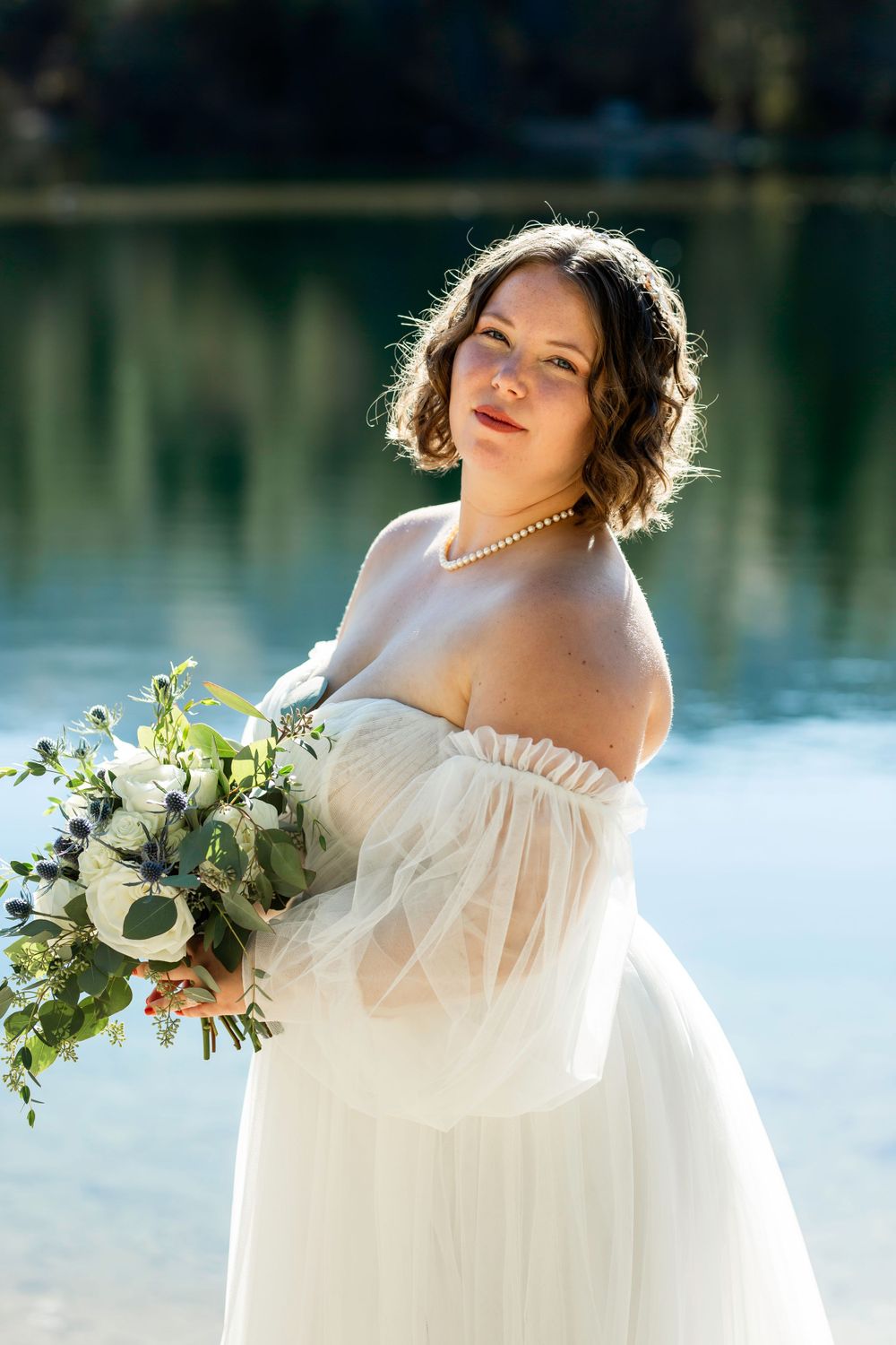 Bride holding white and green bouquet poses by serene lake at sunset.