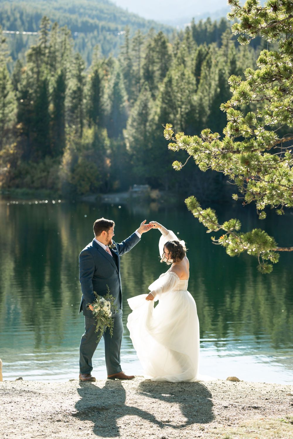 Couple dancing by mountain lake with sunlit pine branches in foreground.