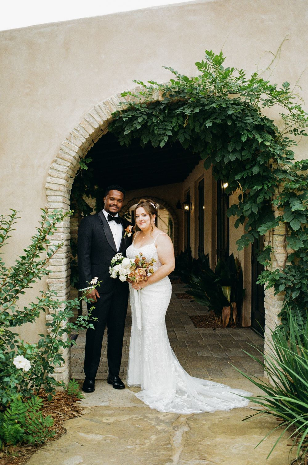 Bride and groom posing under a lush ivy-covered brick arch at an elegant outdoor wedding venue.