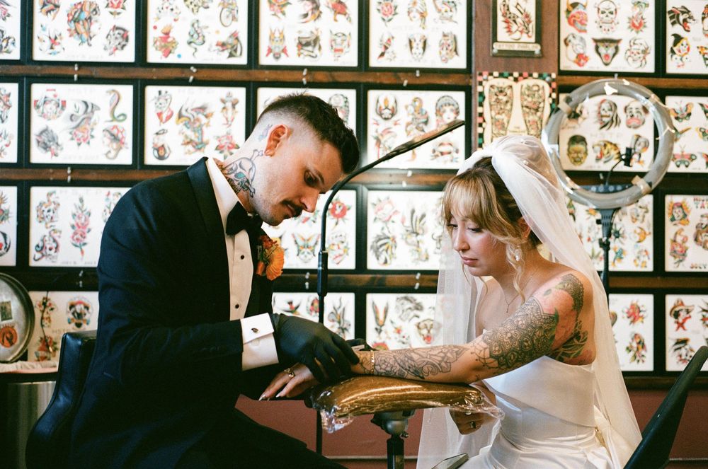 Tattooed bride and groom in wedding attire sitting together in a tattoo parlor surrounded by flash art.