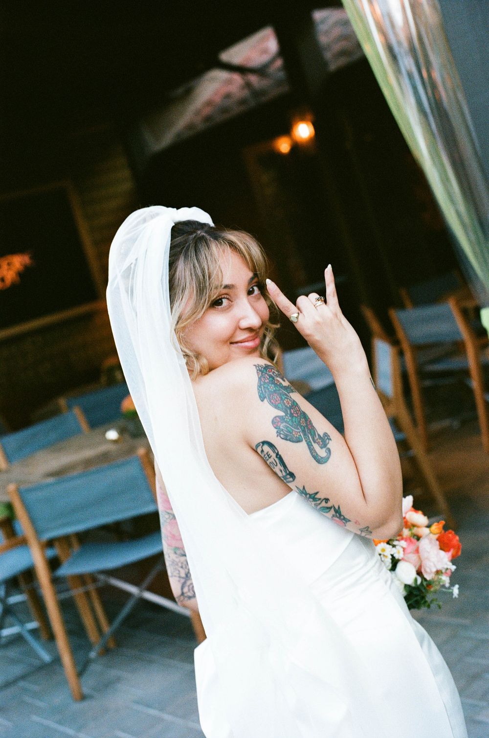 Tattooed bride in white gown and veil making rock sign holding bouquet.