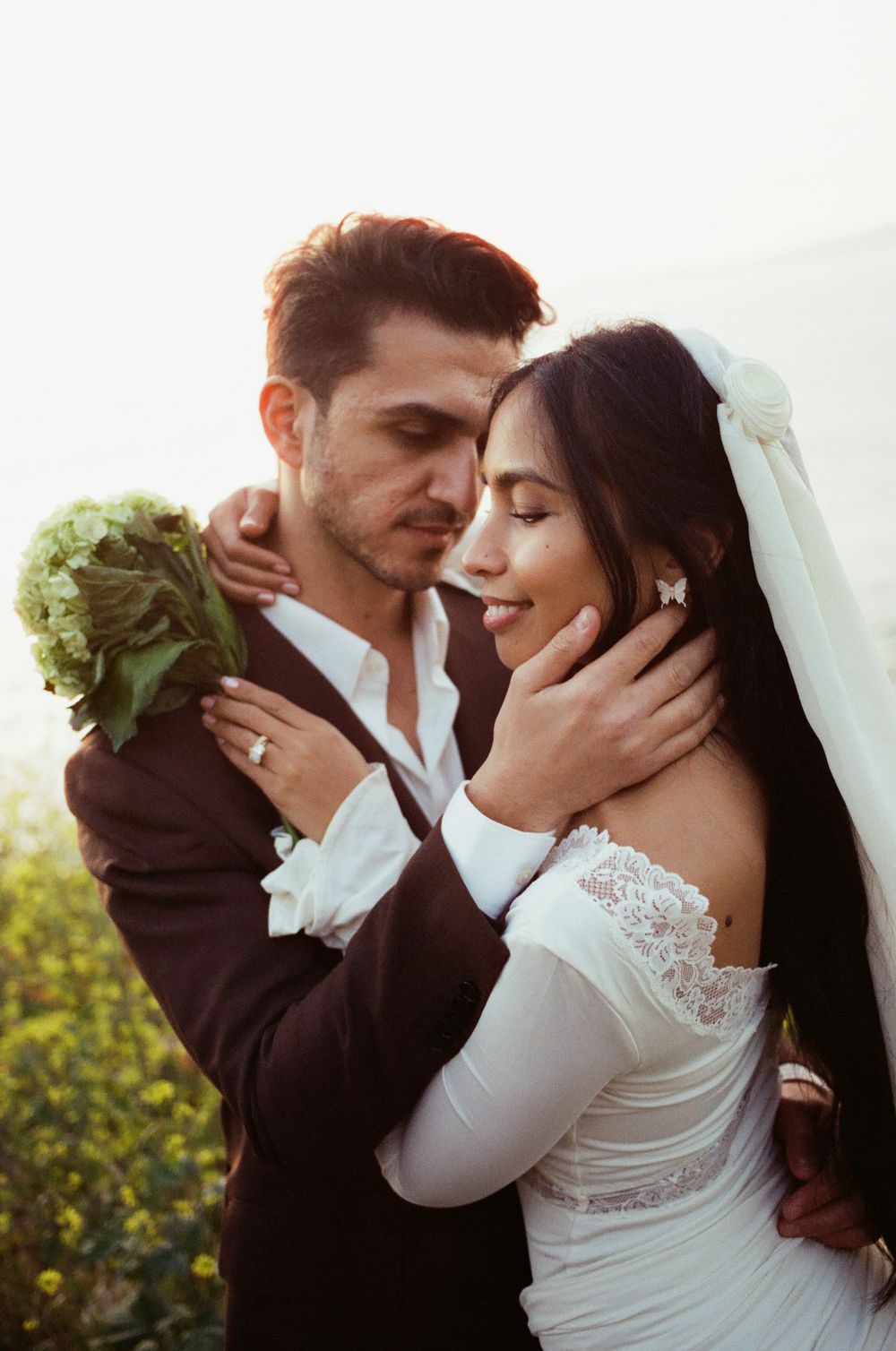 Bride and groom embracing outdoors, bride in lace off-shoulder gown with veil, groom in dark suit holding bouquet.