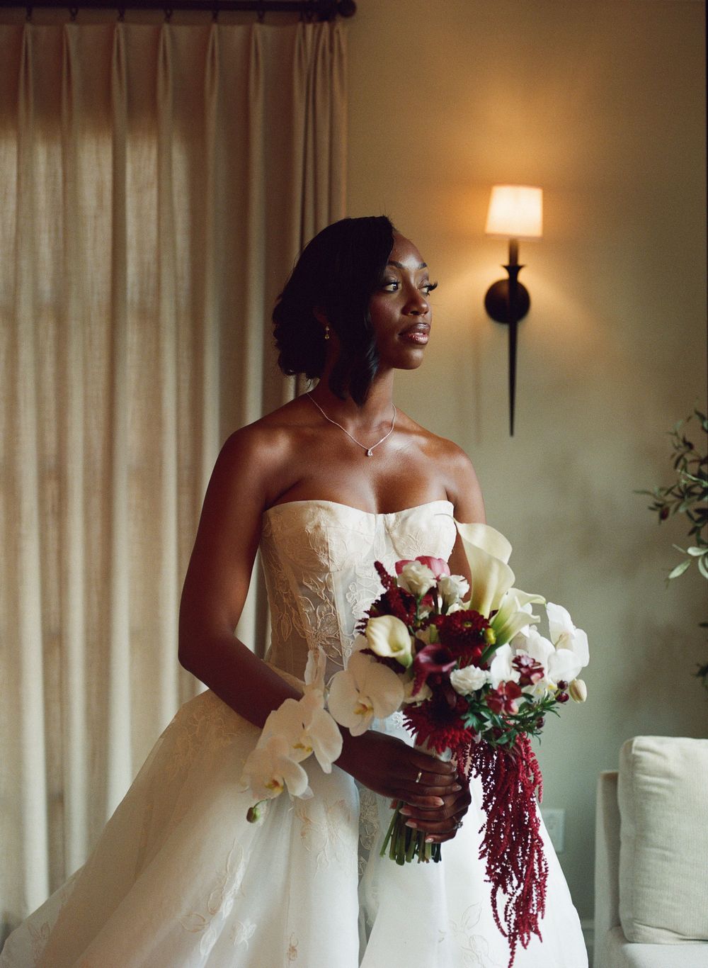 Bride in strapless lace gown holds red and white bouquet by window with warm sconce light.