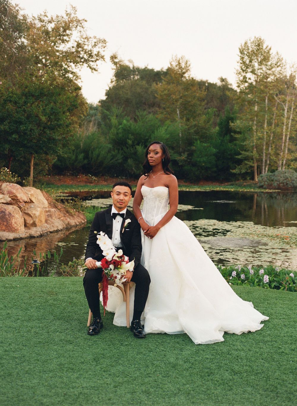 Elegant interracial couple posing in wedding attire beside a serene pond surrounded by lush greenery.