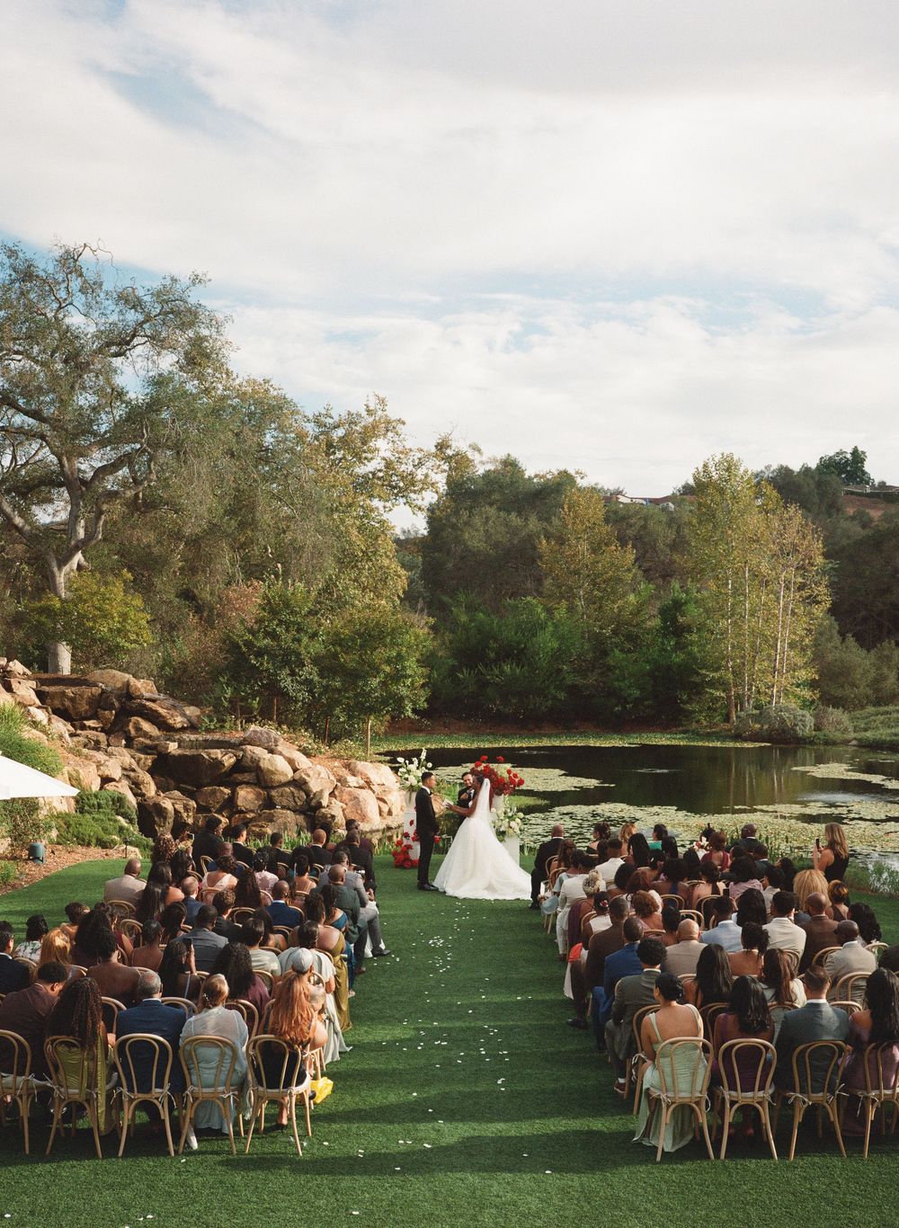 Outdoor wedding ceremony on green lawn with pond and trees, guests seated in rows facing couple at floral arch altar.
