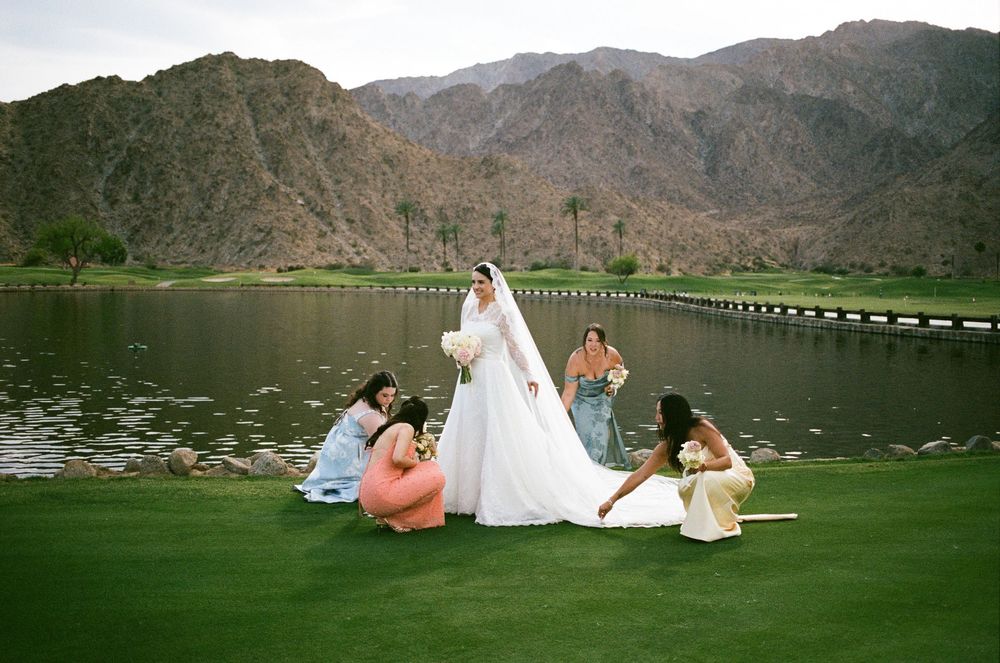 Bride in white gown with veil surrounded by bridesmaids on green golf course with mountain lake backdrop.