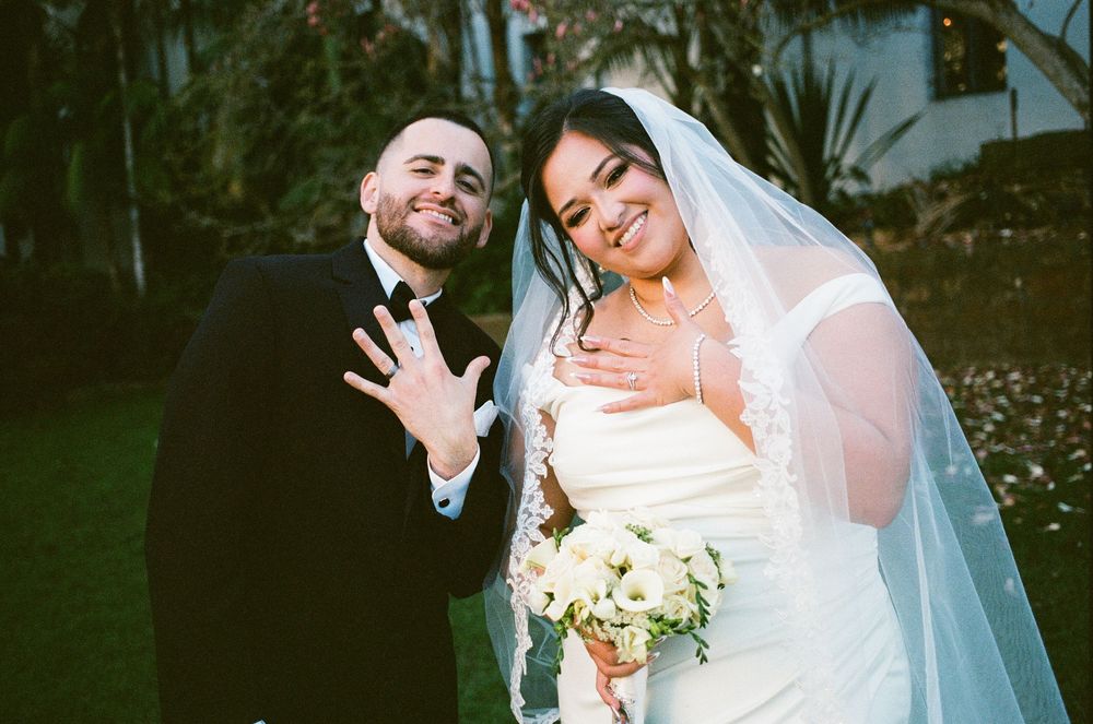 Smiling newlyweds in wedding attire showing off rings, bride holding white bouquet with veil, outdoors.
