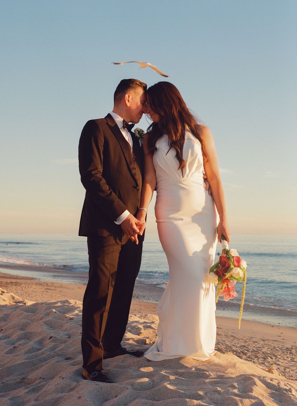 Bride and groom sharing romantic moment on sandy beach at sunset with ocean in background.