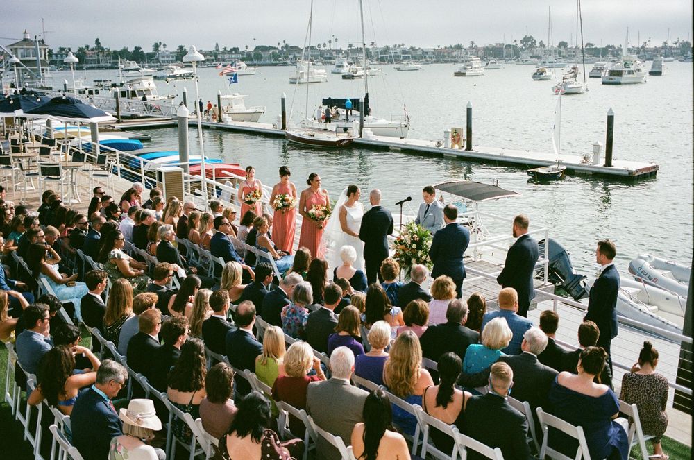 Outdoor waterfront wedding ceremony with guests seated in white chairs beside a marina with sailboats in the background.