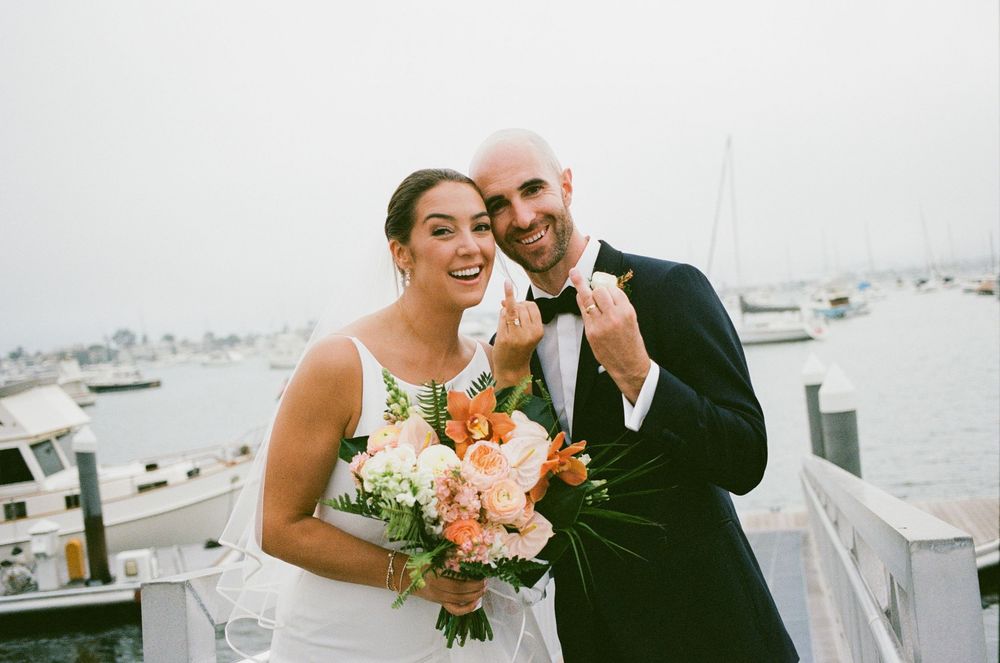 Happy newlyweds pose with orange floral bouquet on waterfront dock with marina and sailboats in background.