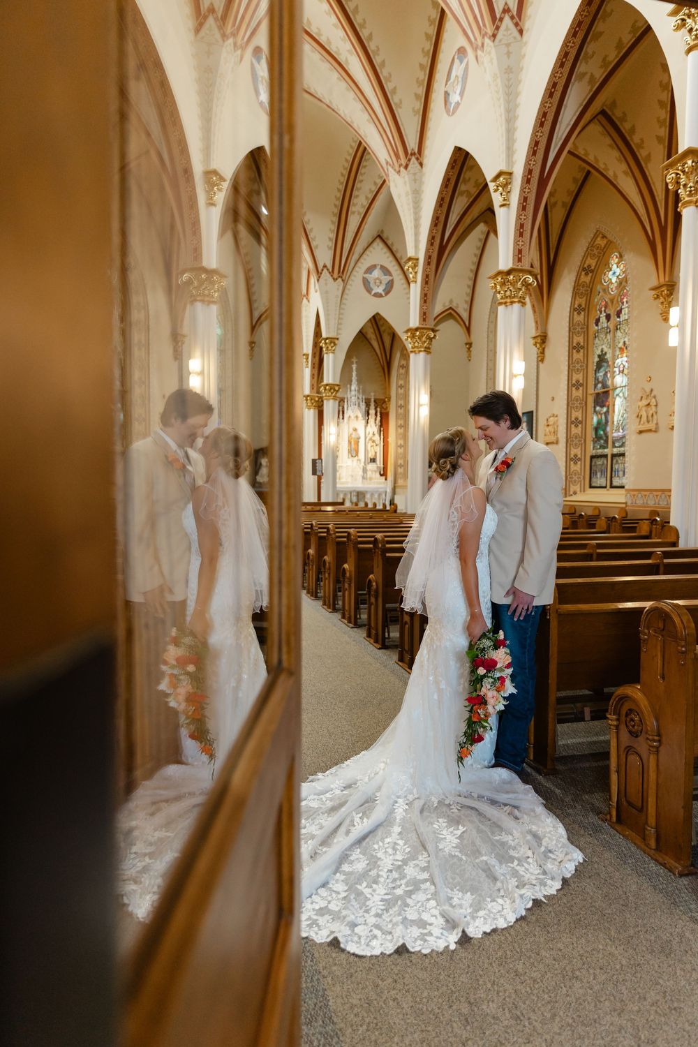 Bride and groom share a kiss in an ornate church aisle, reflected in a wooden door, bride in lace gown with floral bouquet.