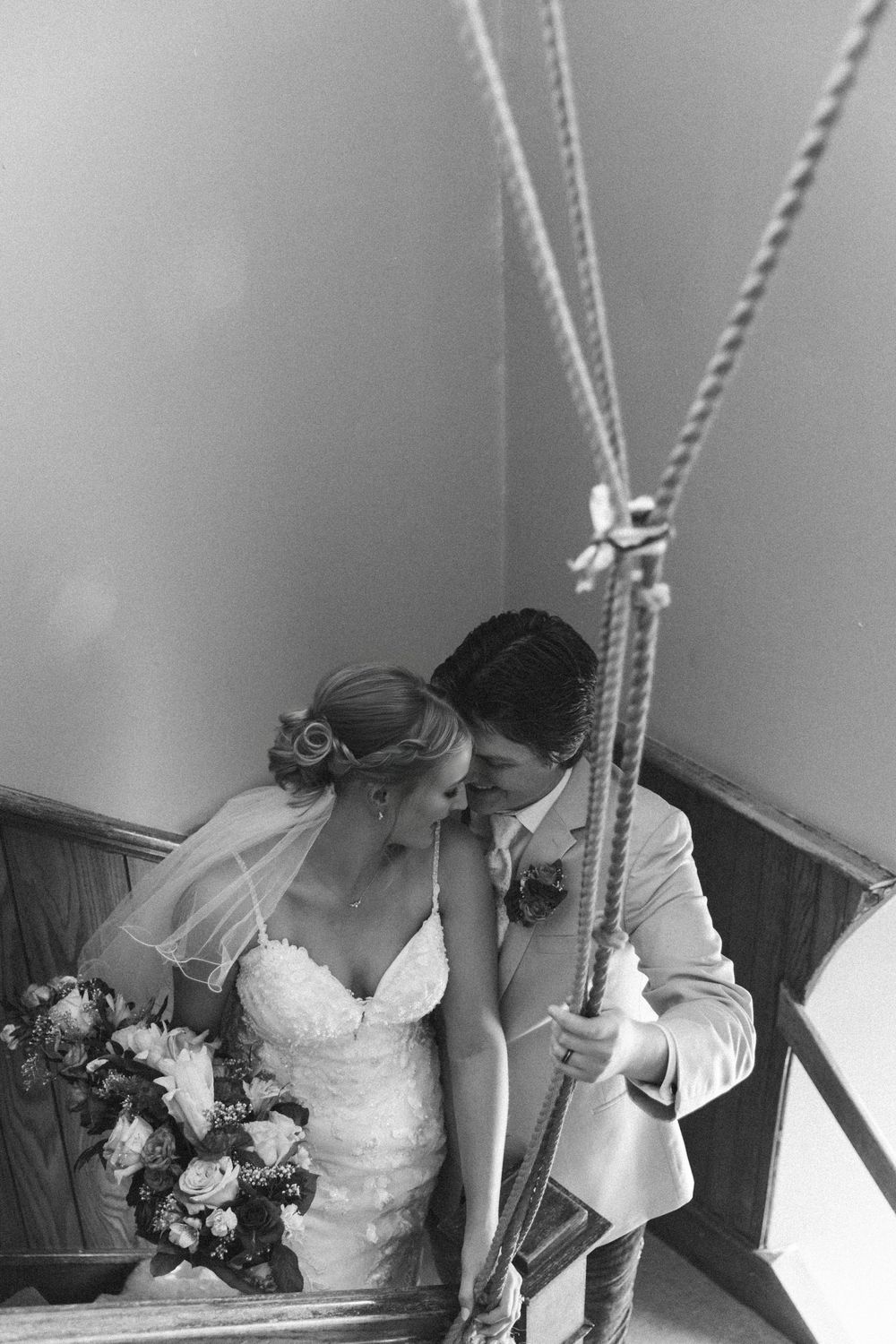 Black and white photo of a bride and groom sharing an intimate moment on a staircase, bride holding a floral bouquet.