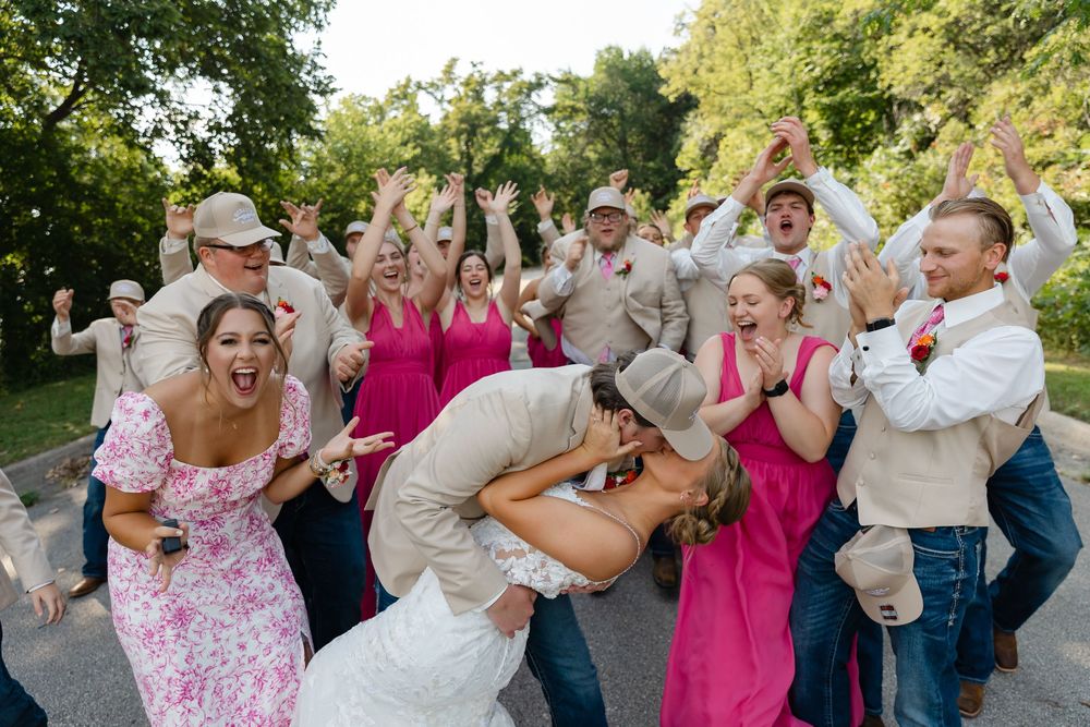 Wedding couple shares a dip kiss while bridal party in pink and tan celebrates joyfully outdoors.