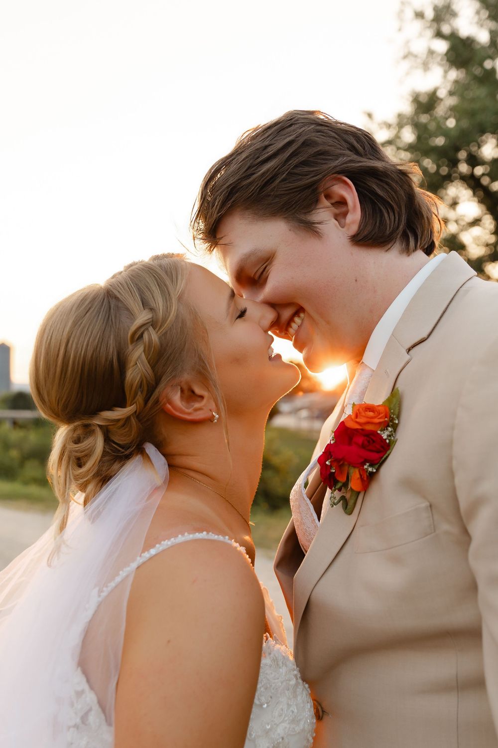 A bride and groom share a romantic almost-kiss at sunset, glowing light between them, she in white lace, he in a tan suit.
