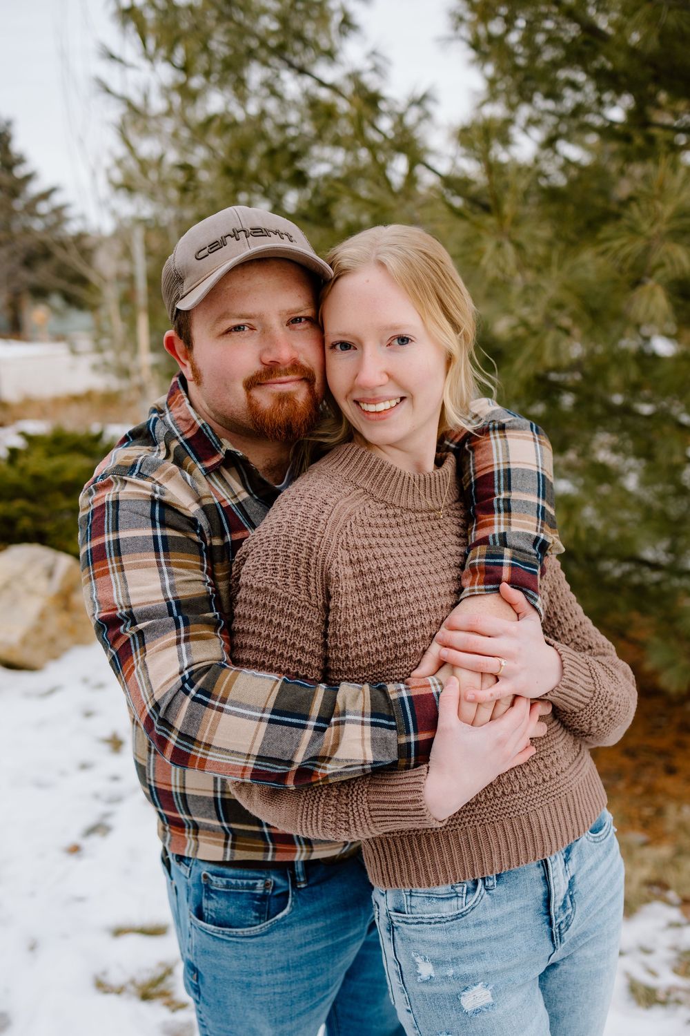 Happy couple embracing outdoors in winter, wearing flannel shirts and a brown sweater, smiling in a snowy evergreen setting.