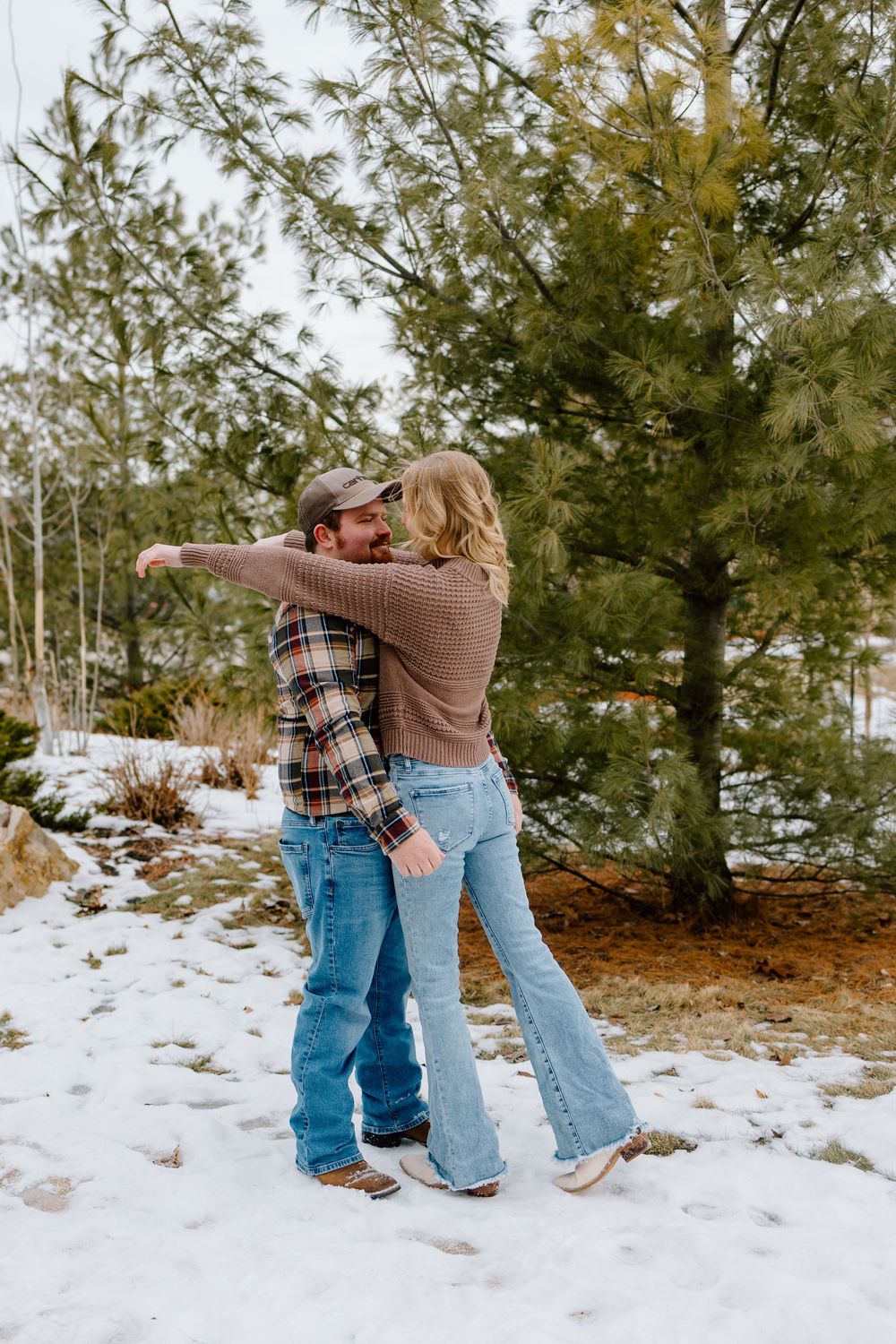 Couple embracing and dancing in snow near evergreen trees, wearing jeans and flannel in a winter outdoor setting.