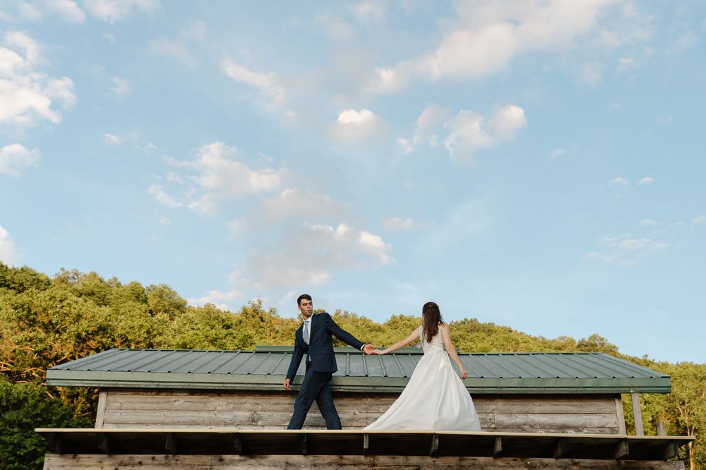 Bride and groom hold hands on a rustic rooftop with lush green trees and blue sky backdrop.