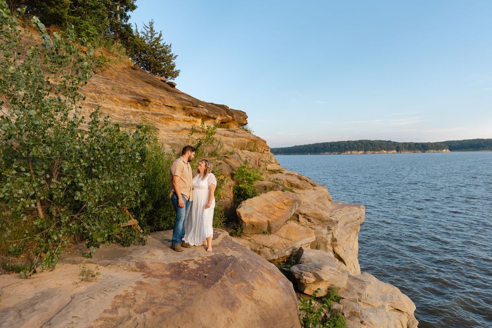 Couple standing on rocky cliffs overlooking a serene lake at golden hour, surrounded by lush greenery and sandstone formations.