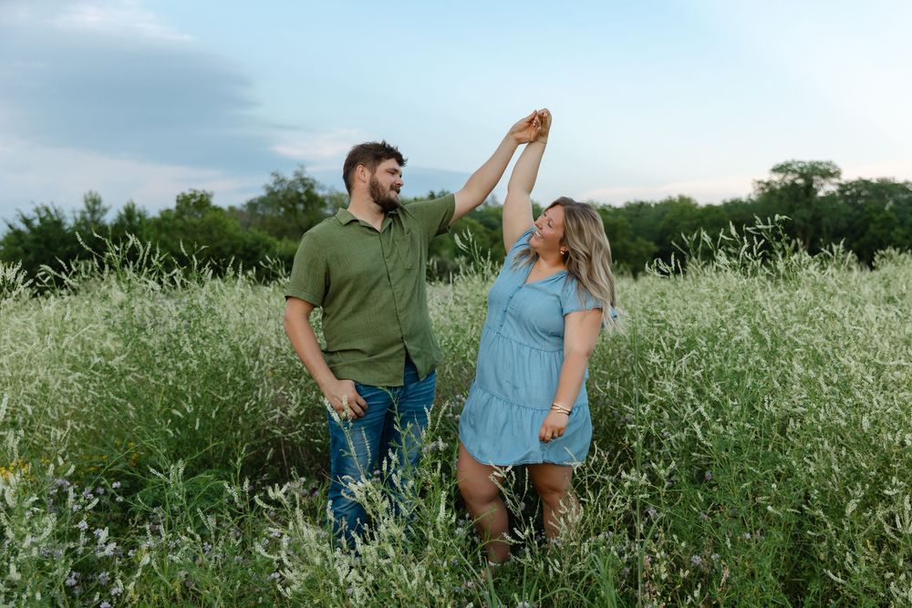 Couple dancing together in a lush green field at dusk, man in green shirt spinning woman in blue dress.