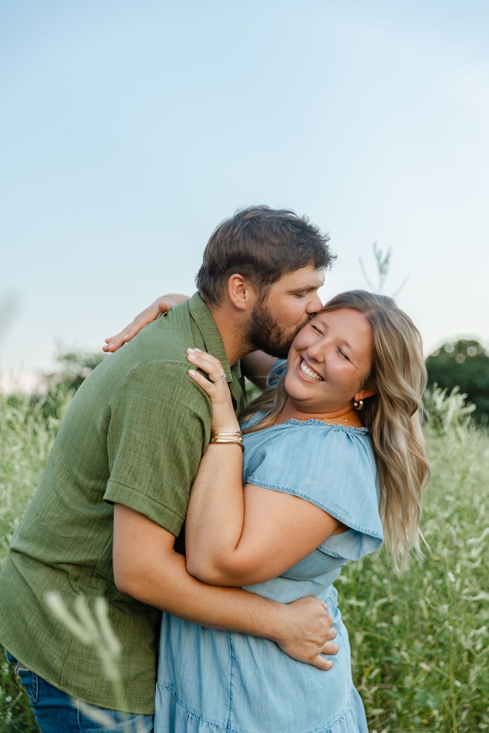 A couple embraces in a green field, the man kissing the woman's cheek as she smiles joyfully.