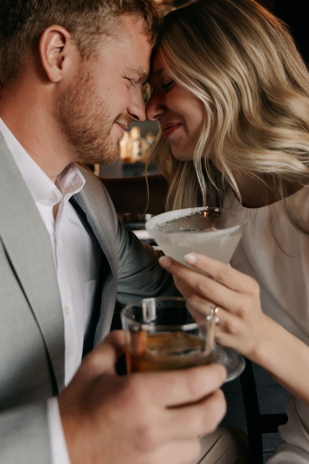 A romantic couple in formal attire clink cocktail glasses, touching foreheads and smiling intimately.