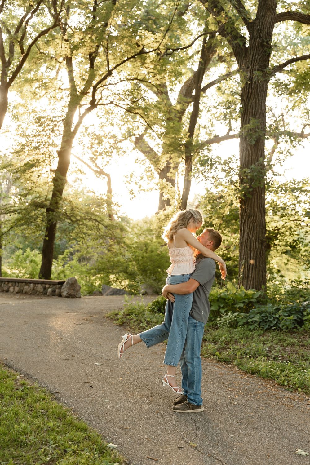 Couple sharing a romantic lift and kiss on a sunlit park path surrounded by lush green trees.