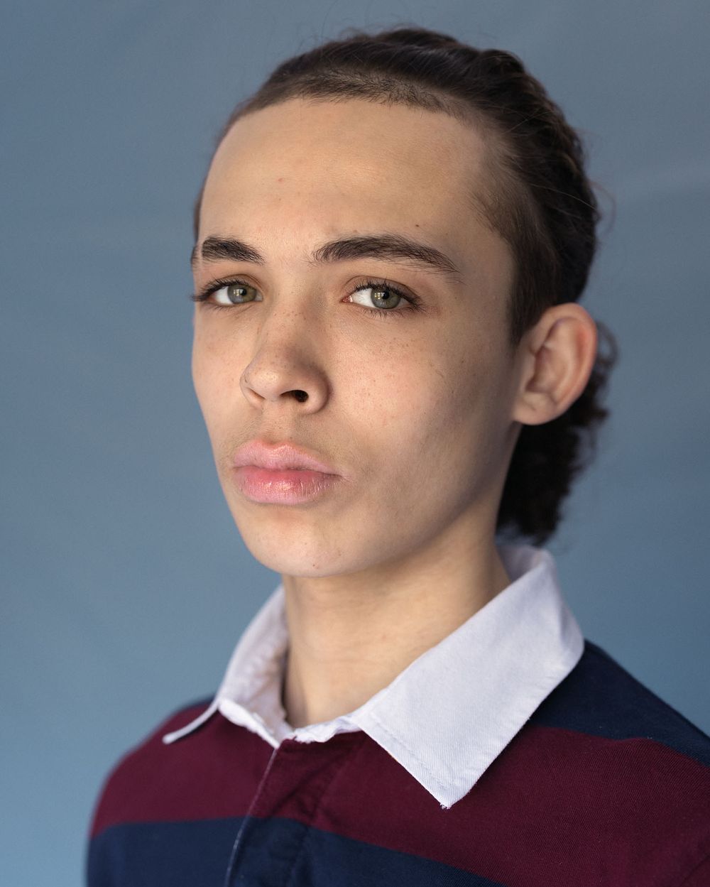 Young male actor portrait wearing burgundy sweater with white collar against blue-grey background for theatrical headshot