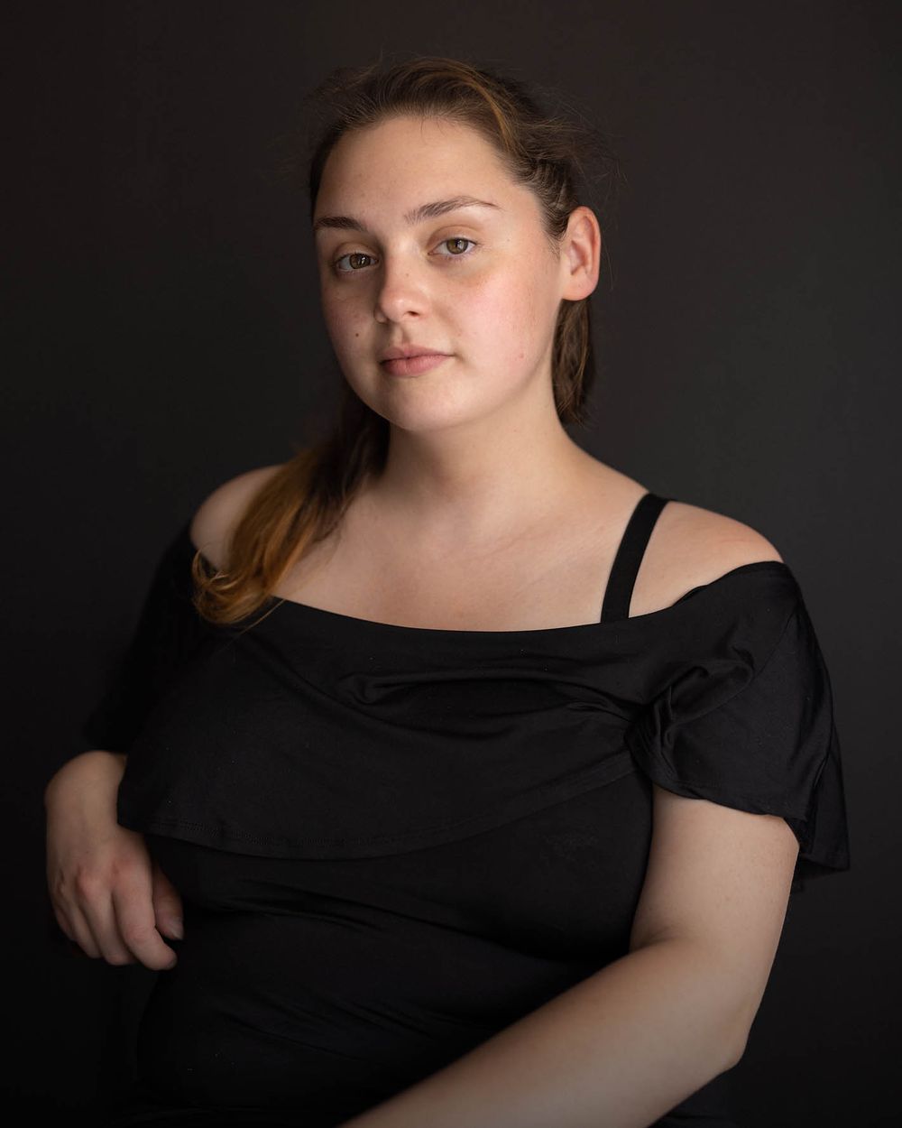Young actress headshot wearing black off-shoulder top against dark background for theatrical portfolio