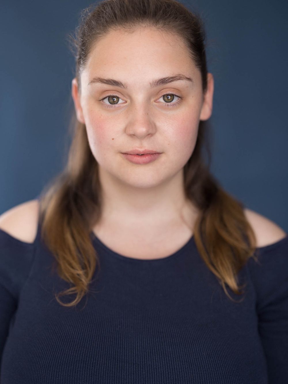 Young woman actor portrait with hair pulled back wearing navy top against blue backdrop for commercial headshot