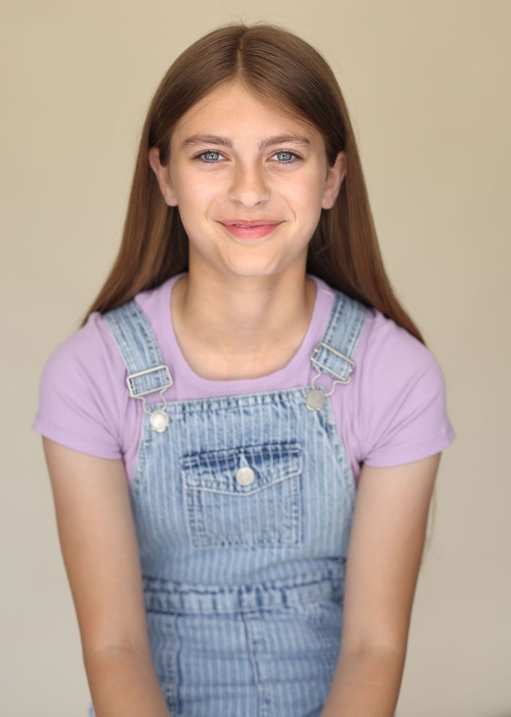 Young girl with long brown hair wearing lavender t-shirt under denim overalls against cream background for commercial child headshot