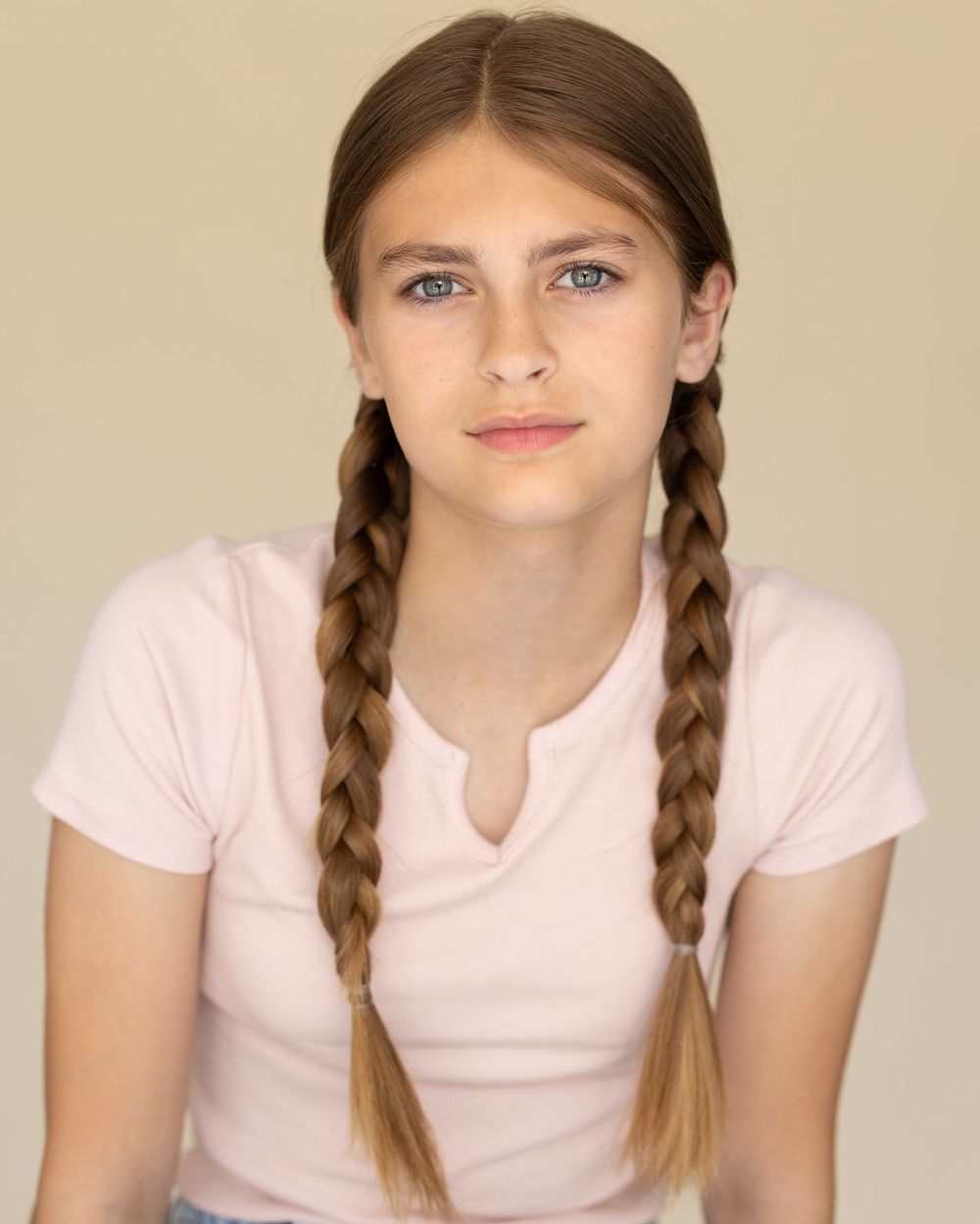 Teenage girl actor headshot with long braided hair wearing pink t-shirt against cream backdrop for commercial portfolio