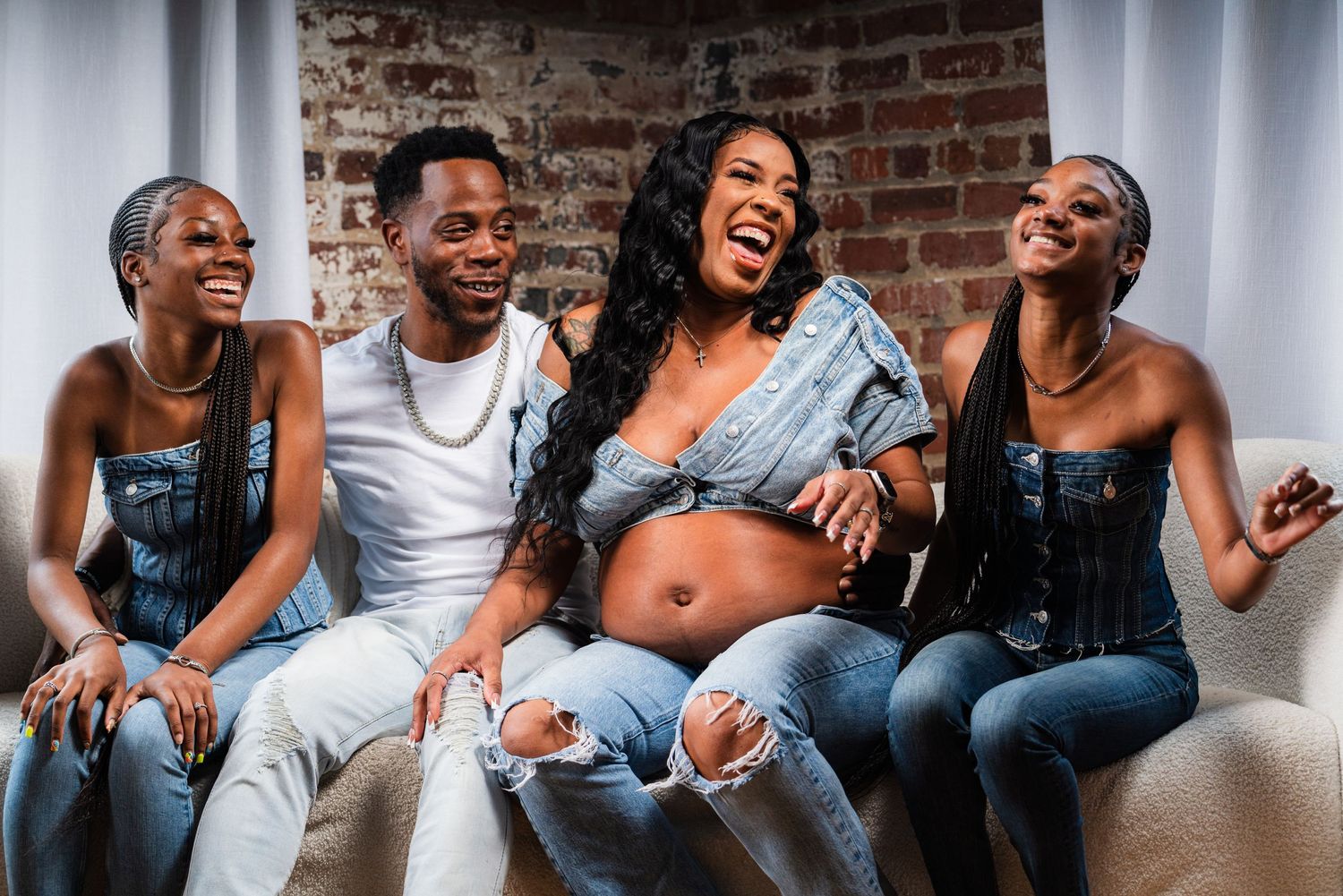 Group laughing together on couch wearing denim against brick wall background.