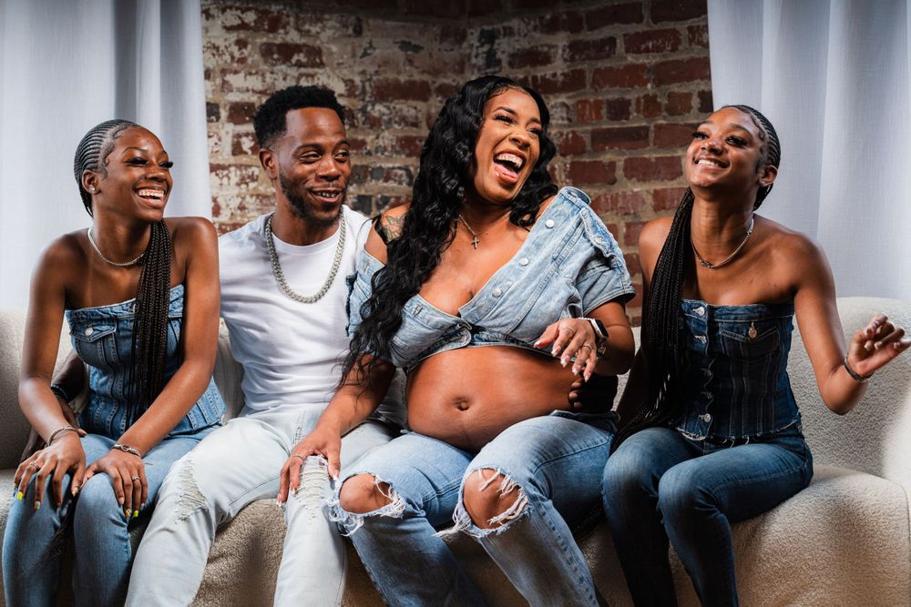 Group laughing together on couch wearing denim against brick wall background.