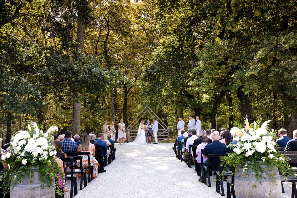 An outdoor wedding ceremony setup with white flower arrangements along a tree-lined path filled with guests on wooden chairs.