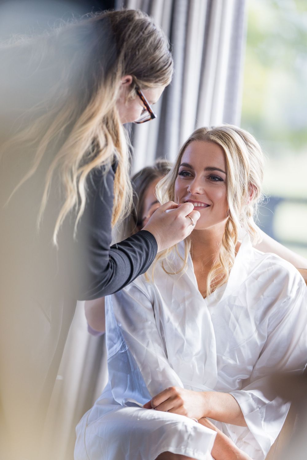 A makeup artist applies finishing touches to a model wearing a white silk robe during a bridal preparation session.