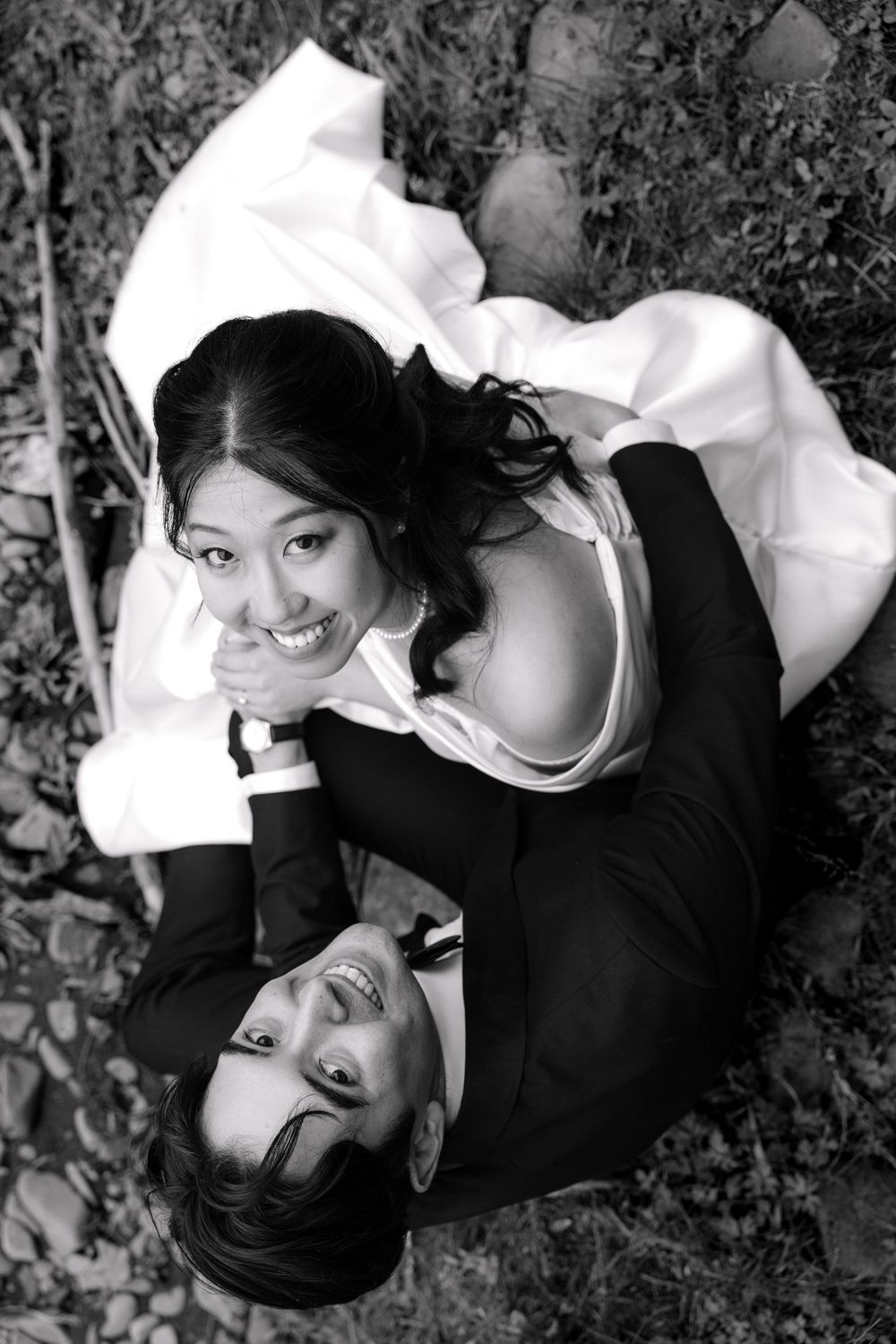 A black and white portrait photograph shows a couple lying on grass looking up at the camera with joyful expressions.