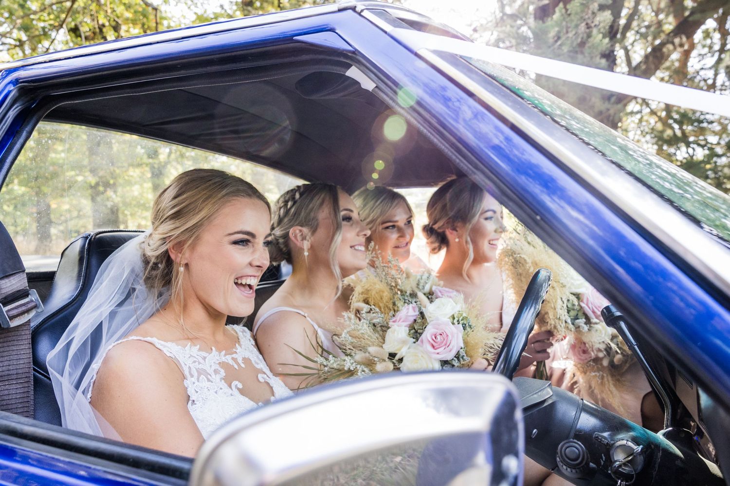 A joyful group sits in a bright blue car, with wedding flowers and the bride's veil visible in sunlight.