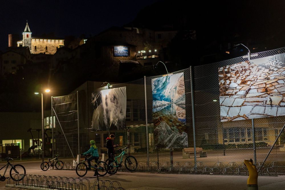 Cyclists ride past illuminated billboards at night with a hilltop church glowing in the background.