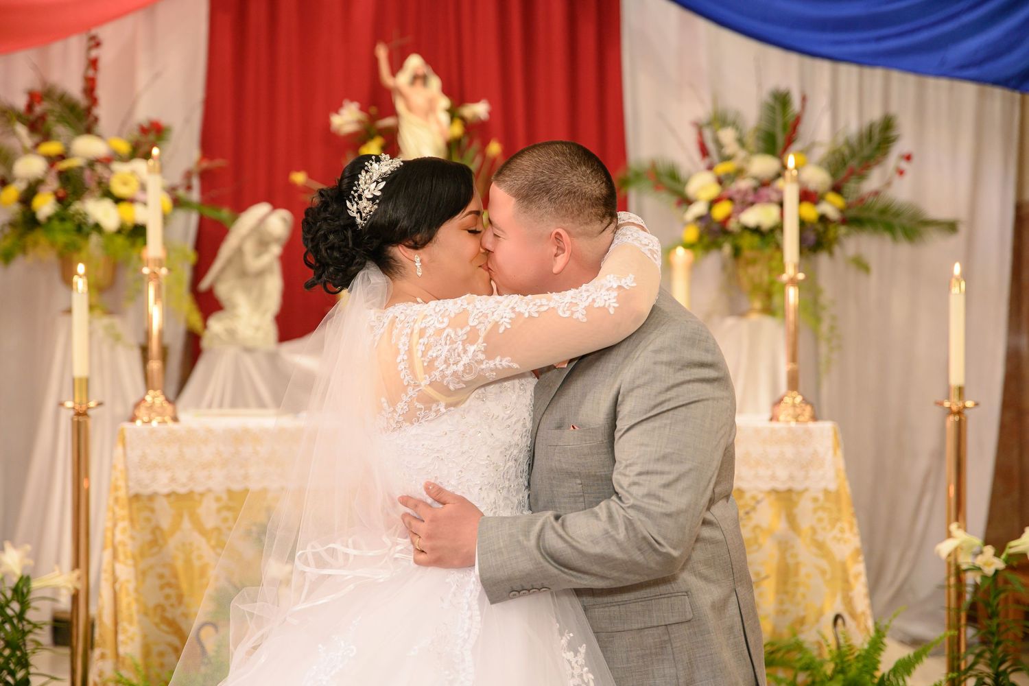 A romantic wedding kiss between newlyweds in front of an elegant white and blue backdrop with floral decorations.